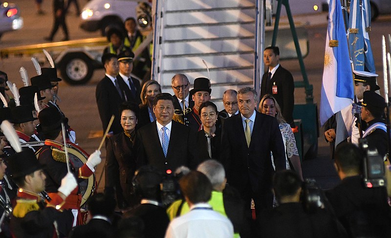 A group of individuals, including Xi Jinping, step off a staircase from an airplane, as a band plays to the left and flag bearers wave to the right.